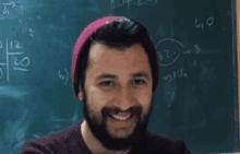 a man with a beard wearing a pink hat is smiling in front of a blackboard with math problems on it .