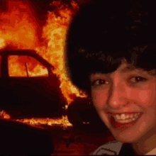 a woman is smiling in front of a burning car in the background .