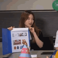 a woman is sitting at a table holding a blue folder with a picture of a cake on it .