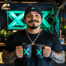 a man wearing a black shirt and a beanie is holding two coffee mugs with x on them