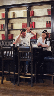 a man and a woman sit at a table in front of a bookcase