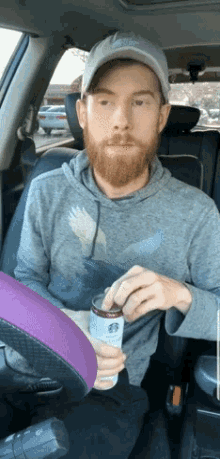 a man with a beard is sitting in the driver 's seat of a car holding a can of starbucks coffee