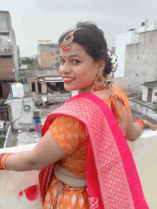 a woman wearing an orange top and a pink saree stands on a balcony