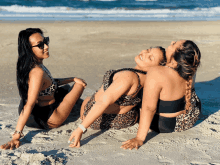 three women in leopard print bikinis are sitting on a sandy beach