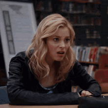 a woman in a leather jacket is sitting at a desk in front of a bookshelf in a library .