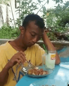 a man in a yellow shirt is sitting at a table eating food and drinking a cup of pocari sweat .