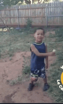 a young boy in a blue tank top and shorts is dancing in the dirt in front of a wooden fence