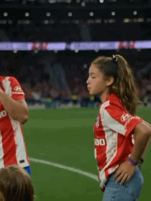 a girl in a red and white hyundai jersey stands on a soccer field with her hands on her hips