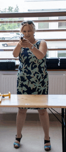 a woman in a floral dress is holding a gun in front of a window