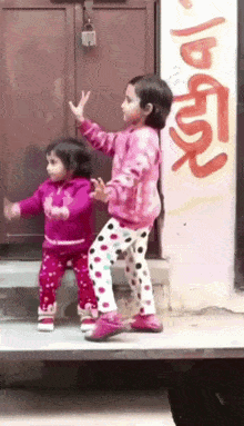 two little girls are dancing in front of a building with graffiti on the wall that says ' a '