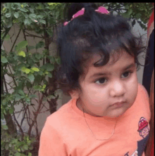 a little girl wearing an orange shirt and a necklace is looking at the camera .