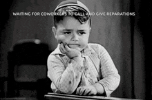 a black and white photo of a little boy sitting at a desk with his hand on his chin .