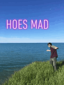 a man is standing on a cliff overlooking the ocean with the words `` hoes mad '' above him .