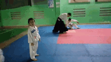 a little girl in a karate uniform is standing on a mat in a gym watching a man do a karate move