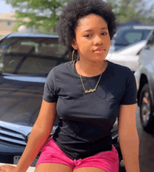 a woman wearing a black shirt and pink shorts is sitting in front of a car with the name matiya on her necklace