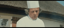 a man in a chef 's hat is standing in front of a thatched building .