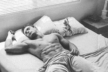 a shirtless man is laying on a bed with a towel around his waist and a window in the background .