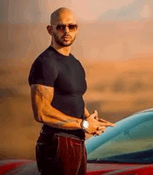 a bald man wearing sunglasses and a black shirt is standing in front of a car .