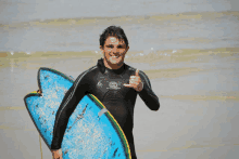 a man in a wetsuit holds a surfboard and gives a thumbs up