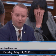 a man in a suit and tie is sitting next to a woman who is covering her mouth during a meeting on may 14th 2019