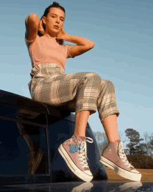 a woman in plaid pants sits on the roof of a car