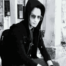 a man wearing a hoodie and sunglasses is sitting on a bench .