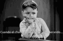 a black and white photo of a little boy sitting at a desk with his hand on his face .