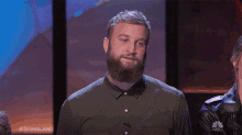 a man with a beard is covering his mouth with his hand while a woman laughs in front of a nbc sign