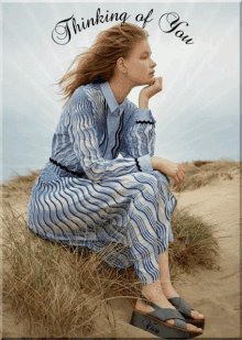 a woman in a blue and white dress is sitting on a sand dune with the words thinking of you above her