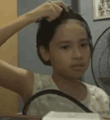 a young girl is adjusting her hair in front of a mirror while sitting in front of a fan .