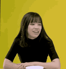 a young woman in a black shirt is sitting at a table with a bowl of food in front of her