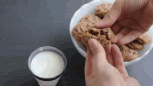 a person is reaching for a chocolate chip cookie from a bowl of cookies next to a glass of milk .