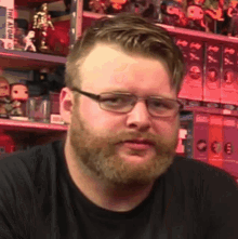 a man with a beard wearing glasses and a black shirt is standing in front of a shelf with toys on it