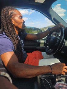 a man with dreadlocks is driving a car and wearing a t-shirt that says d.c. state