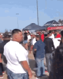 a man in a white shirt is standing in a crowd of people in front of a tent that says tail .