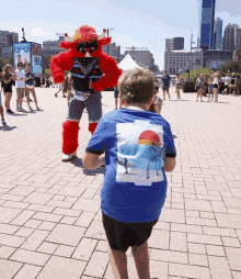 a boy wearing a blue shirt with a picture of a beach on it stands in front of a mascot