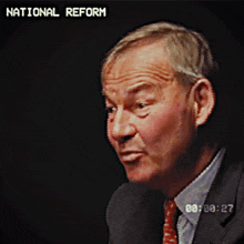 a man in a suit and tie is talking in front of a screen that says " national reform "