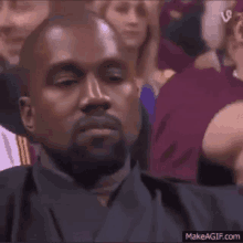 kanye west is sitting in a crowd of people and making a face .