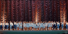 a group of young girls are standing on a stage in front of a large curtain with drop that written on the bottom