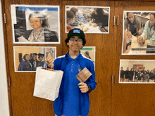 a man in a blue shirt is holding a white bag and a book in front of a wall of pictures .