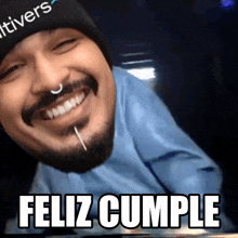 a man wearing a beanie and a blue shirt is smiling and says feliz cumple in white letters