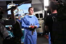 a man in a surgical gown with blood on his hands stands in front of a camera