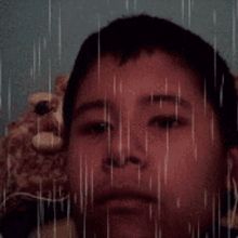 a young boy with a teddy bear in the background looks at the camera with rain drops falling on his face