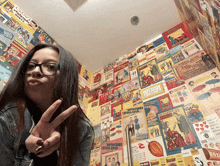 a woman giving a peace sign in front of a wall covered in posters including one that says ' snoopy '