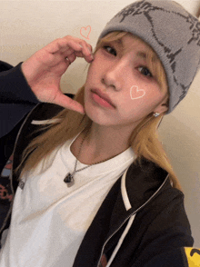 a woman wearing a beanie and a white shirt making a heart shape with her hands