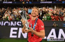 a man in a red shirt is holding a trophy in front of a crowd that is cheering for england