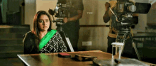 a woman is sitting at a table in front of a camera while a man holds a camera .