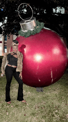 a woman in a fur coat stands next to a large red christmas ornament