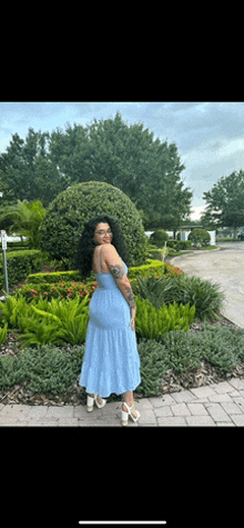 a woman in a blue dress and white heels is standing in front of a garden .