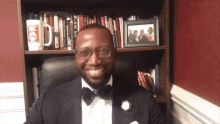 a man in a tuxedo sits in front of a bookshelf with a picture of a man in a suit behind him